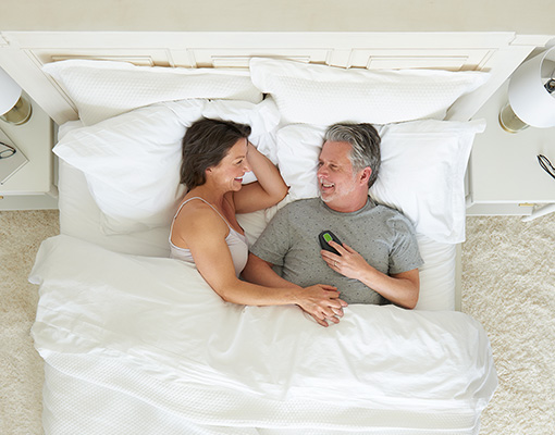 A couple lying in bed seen from above, smiling at each other while he holds the Inspire sleep remote