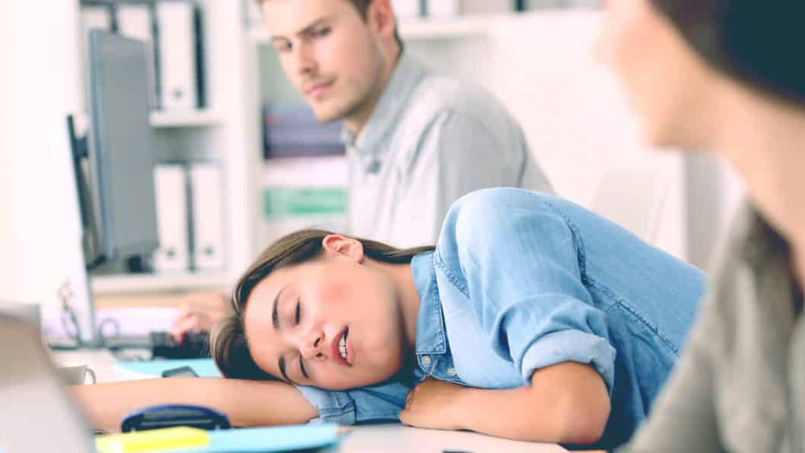A woman asleep at her desk while a colleague works behind her