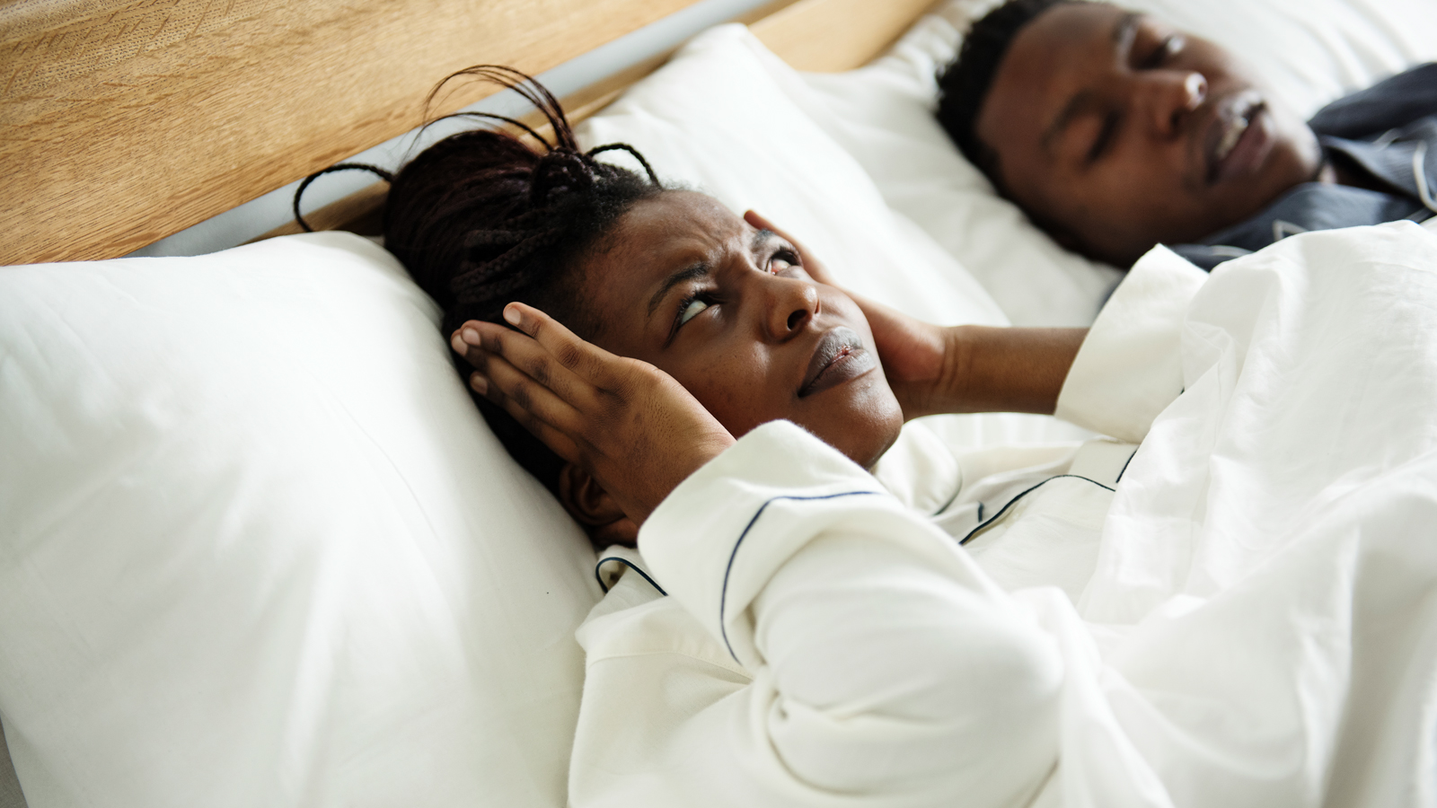 A woman lying awake in bed with her hands over her ears while the man beside her sleeps with his mouth open